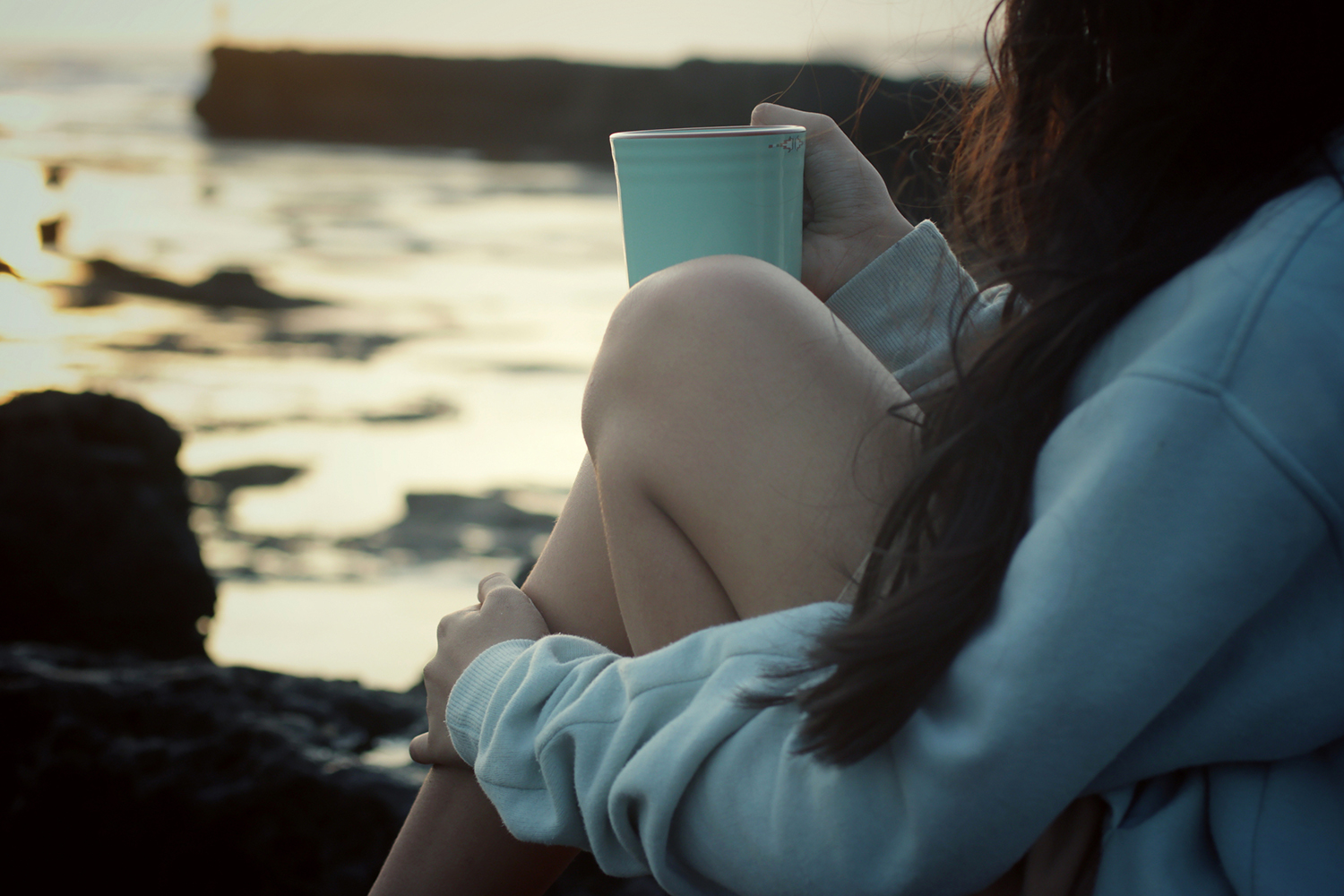 a young woman in comfy clothes drinking tea on a rocky shore