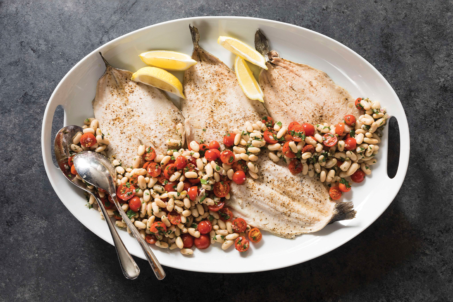 A top view of Roasted Trout with White Bean and Tomato Salad on a white serving tray with a charcoal gray background.