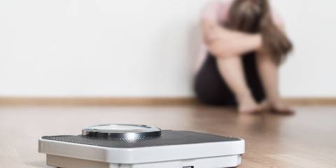 A scale on the floor with a woman sitting in the background with her head hung with failure.