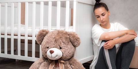 a depressed woman leaning on a crib looking at a teddy bear